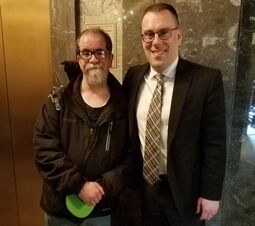 2 Caucasian men stand side by side in front of a elevator at the state capitol. 